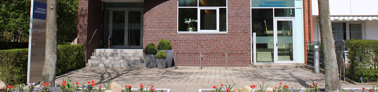 Eingangsbereich eines Backsteingebäudes mit gepflegten Pflanzkübeln, grünen Büschen sowie ein Blumenbeet mit bunten Tulpen.