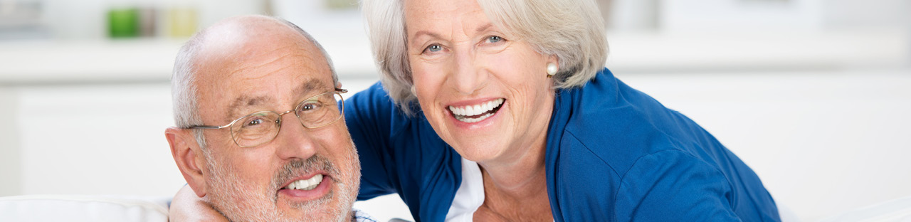 Ein lächelndes älteres Paar, wobei eine Frau mit hellen Haaren in einem blauen Oberteil von hinten einen Mann mit Brille und Bart umarmt und beide frontal in die Kamera blicken.