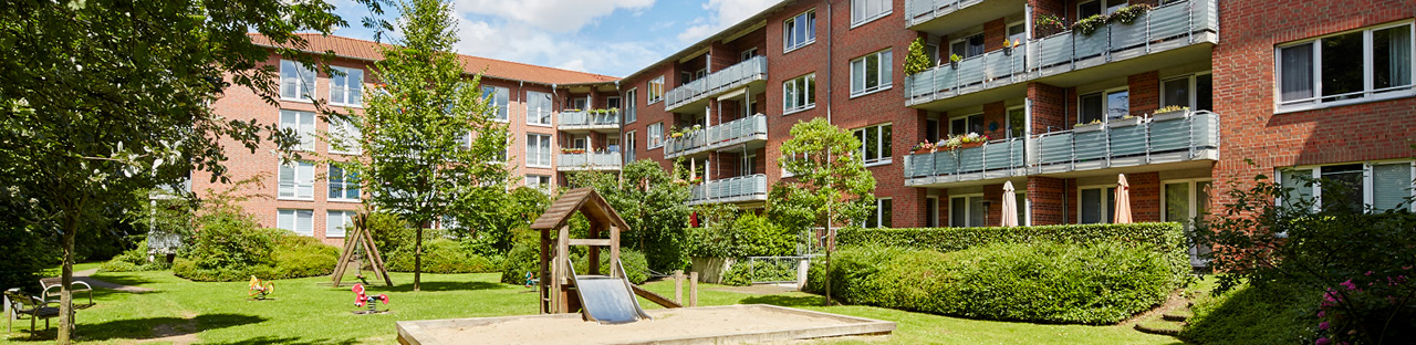 Eine Außenansicht zeigt einen begrünten Innenhof eines U-förmigen, mehrstöckigen Wohnhauses in rotem Backstein mit grauen Balkonen, in dessen Mitte sich ein heller Sandkasten mit einer hölzernen Rutsche und einer Schaukel befindet.